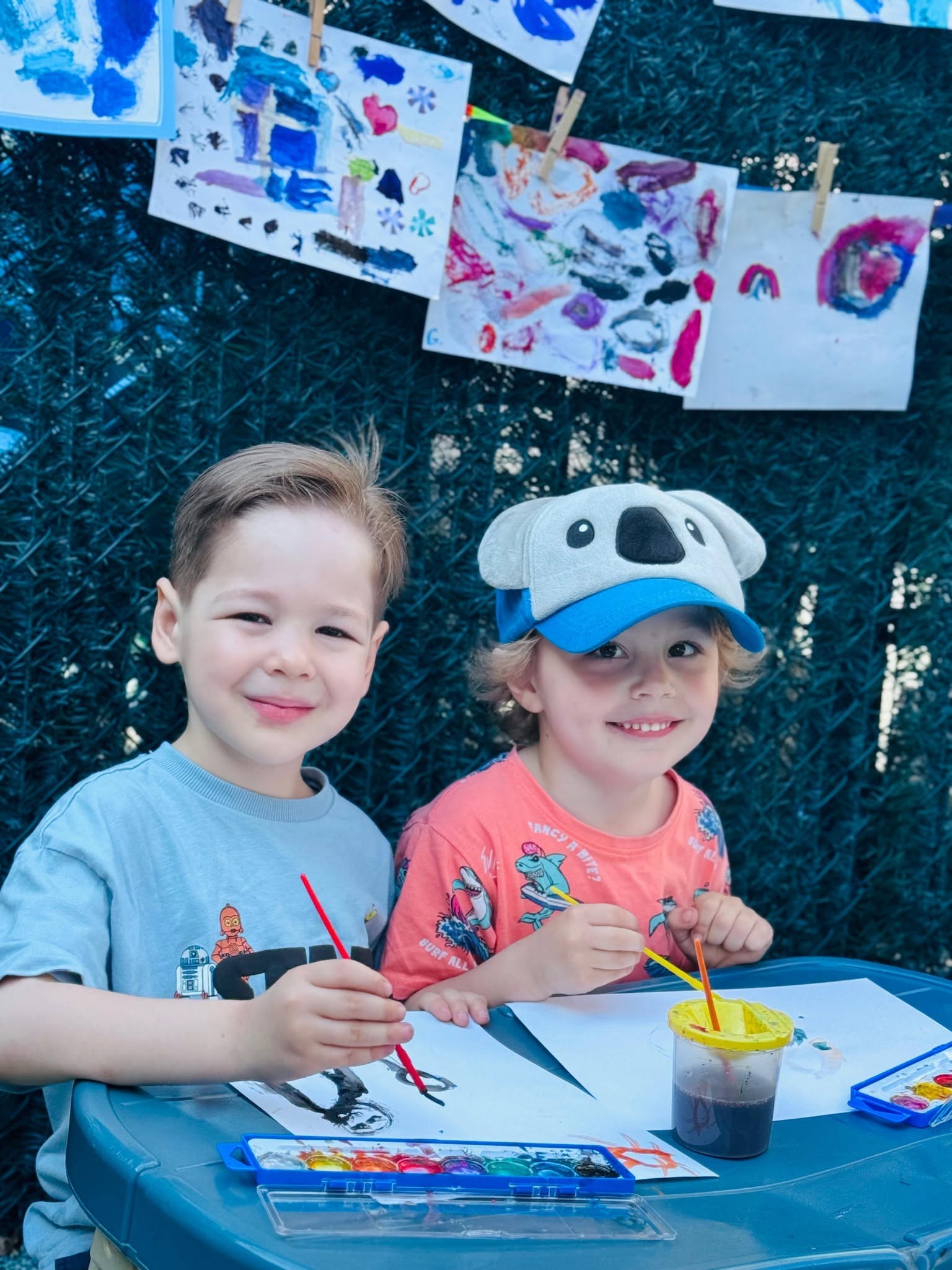 Two smiling boys painting with watercolors at an outdoor table